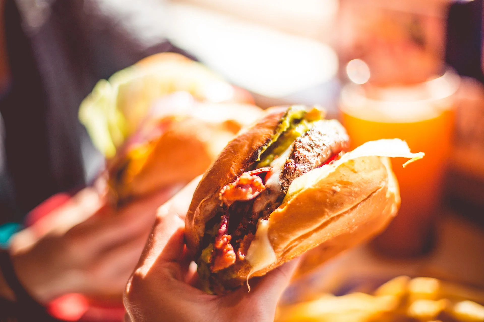 Osoba drži veliki hamburger sa mesom, sirom i povrćem; drugi hamburger i piće su zamućeni u pozadini.