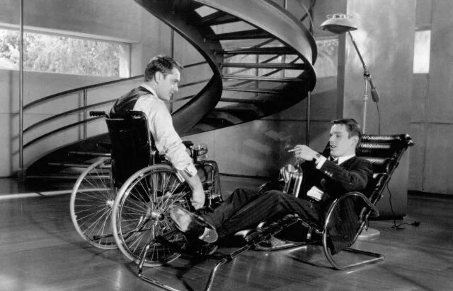 Crno-bela filmska scena sa mu&scaron;karcem u invalidskim kolicima i drugim mu&scaron;karcem u naslonjači, ispred spiralnog stepeni&scaron;ta.