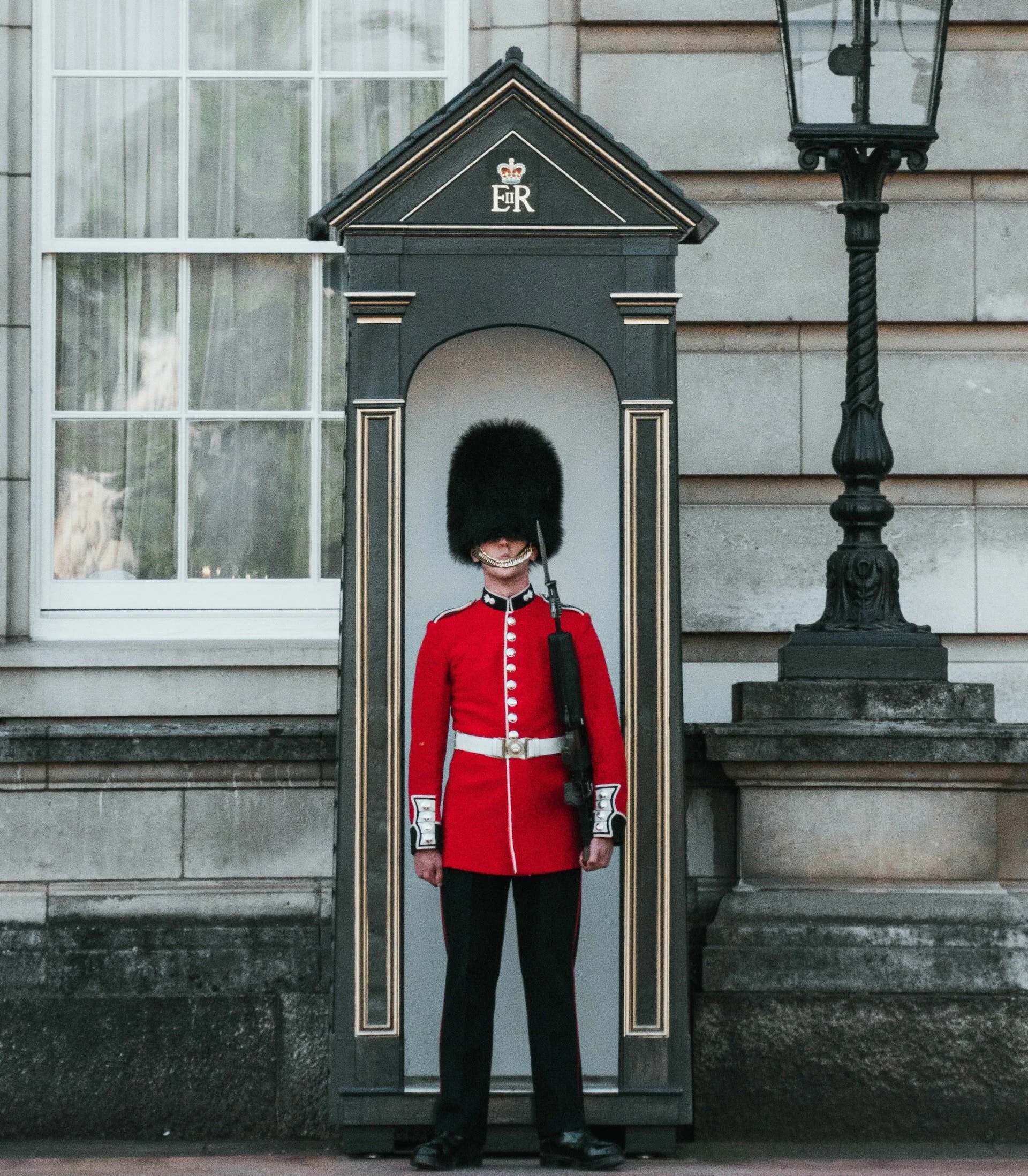 Britanski gardista u crvenoj uniformi i visokoj crnoj kapi stoji mirno u stražarskoj kućici ispred kamene zgrade.