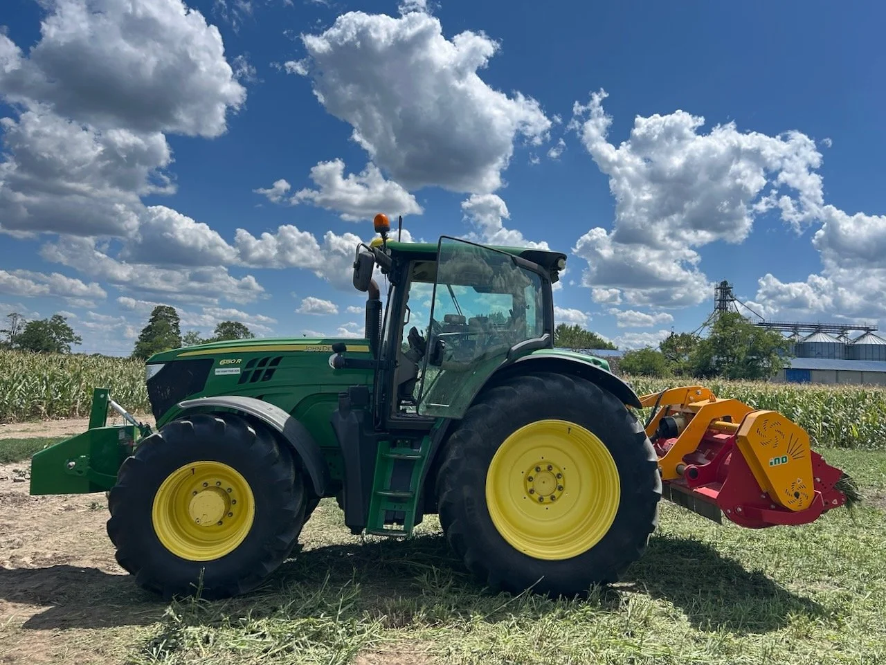 Zeleni John Deere traktor sa crveno-žutim malčerom stoji u polju pod oblacima.