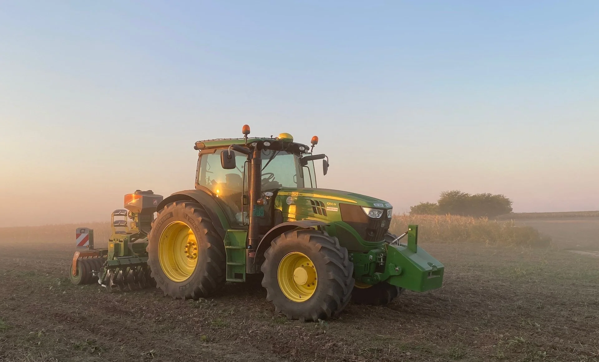 Zeleni traktor sa sejalicom radi na suvom polju u pra&scaron;ini pred zalazak sunca.