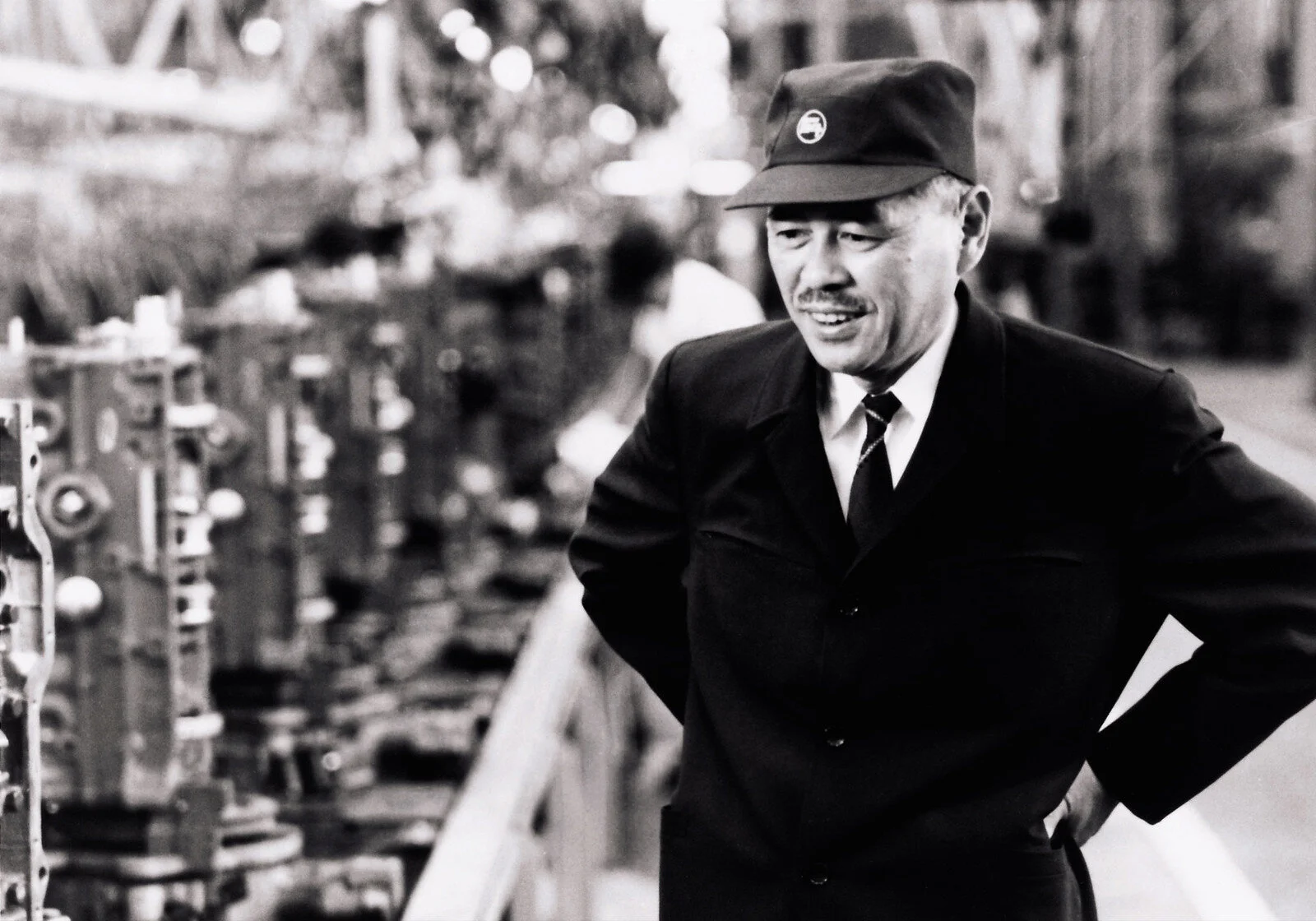 Crno-bela fotografija mu&scaron;karca u industrijskom pogonu, pored proizvodnih ma&scaron;ina.