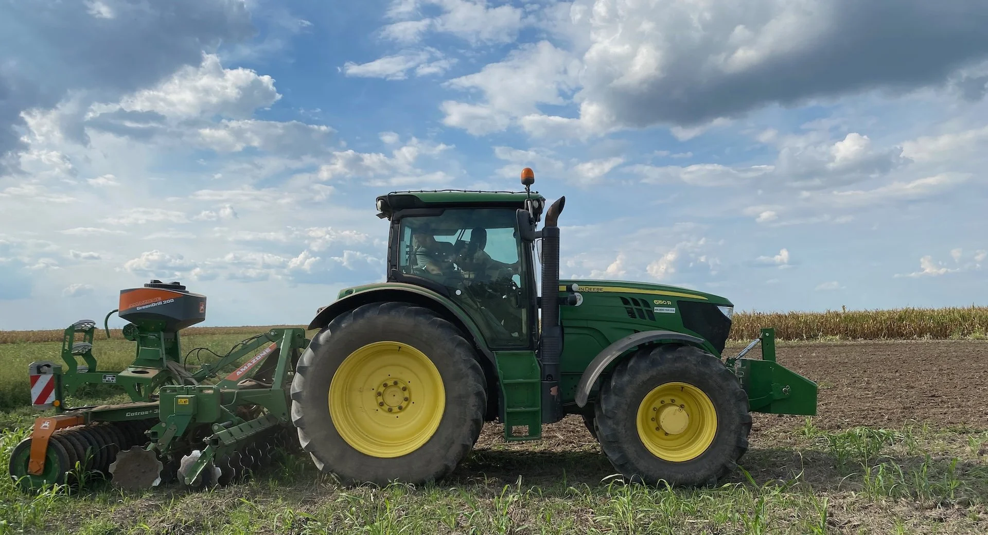 Zeleni traktor sa priključkom radi na njivi pod oblačnim nebom.