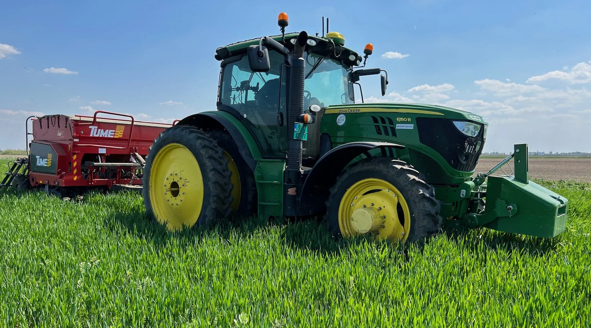 Zeleni traktor sa crvenom sejalicom radi u zelenom polju.