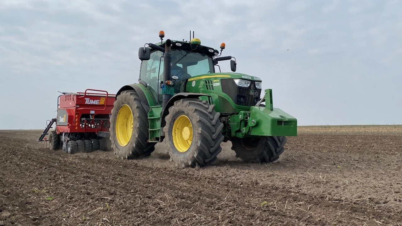 Zeleni traktor sa crvenom sejalicom prelazi preko ogolele, obrađene zemlje.