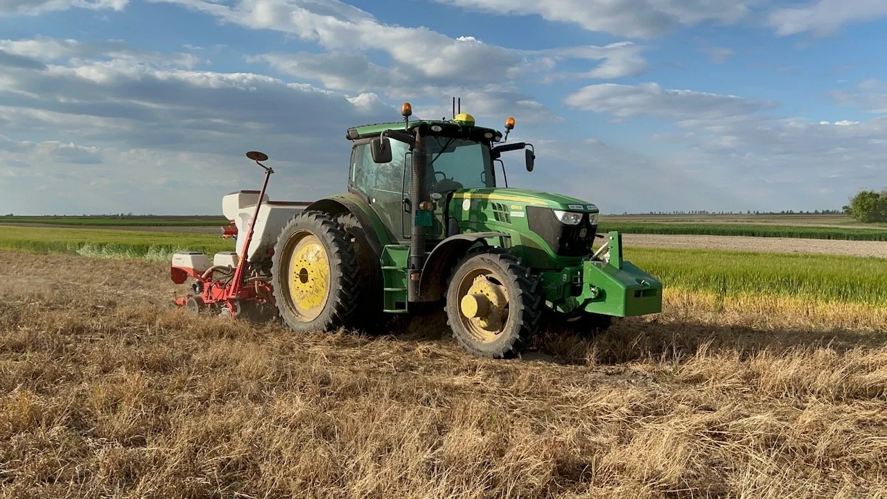 Zeleni traktor sa sejalicom radi kroz strnjiku pod plavim nebom.
