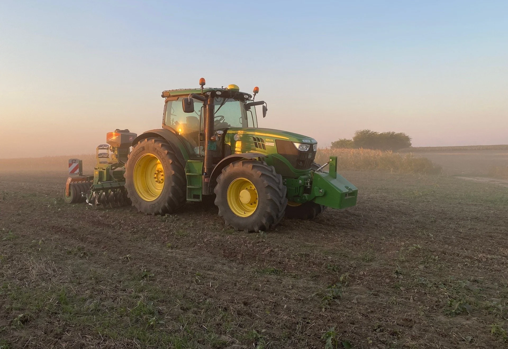 Zeleni traktor sa sejalicom radi na polju obasjanom zalaskom sunca.